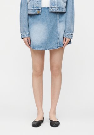 Moteris dėvinti šviesiai mėlyną džinsinę mini sijoną, derančią džinsinę striukę ir juodus plokščius batelius, stovinti prieš baltą foną.