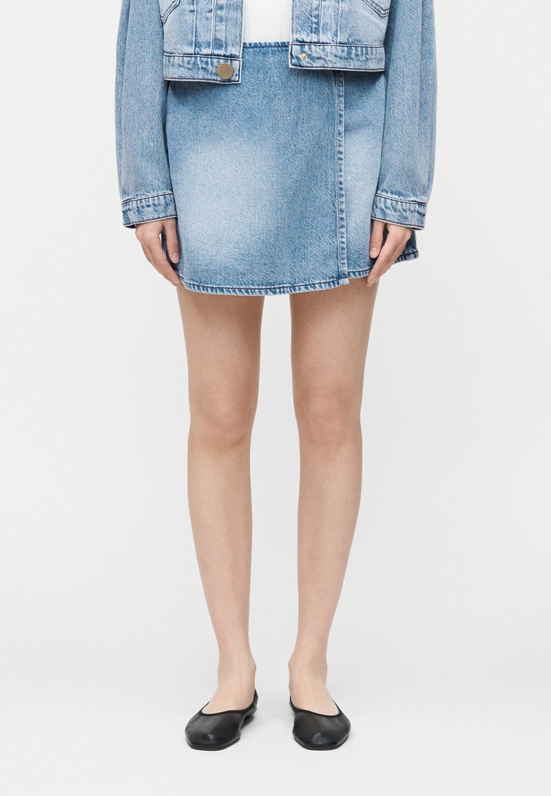 Γυναίκα που φοράει ανοιχτό μπλε μίνι φούστα από τζιν, τζιν μπουφάν στο ίδιο χρώμα και μαύρα επίπεδα παπούτσια, στέκεται μπροστά σε λευκό φόντο.