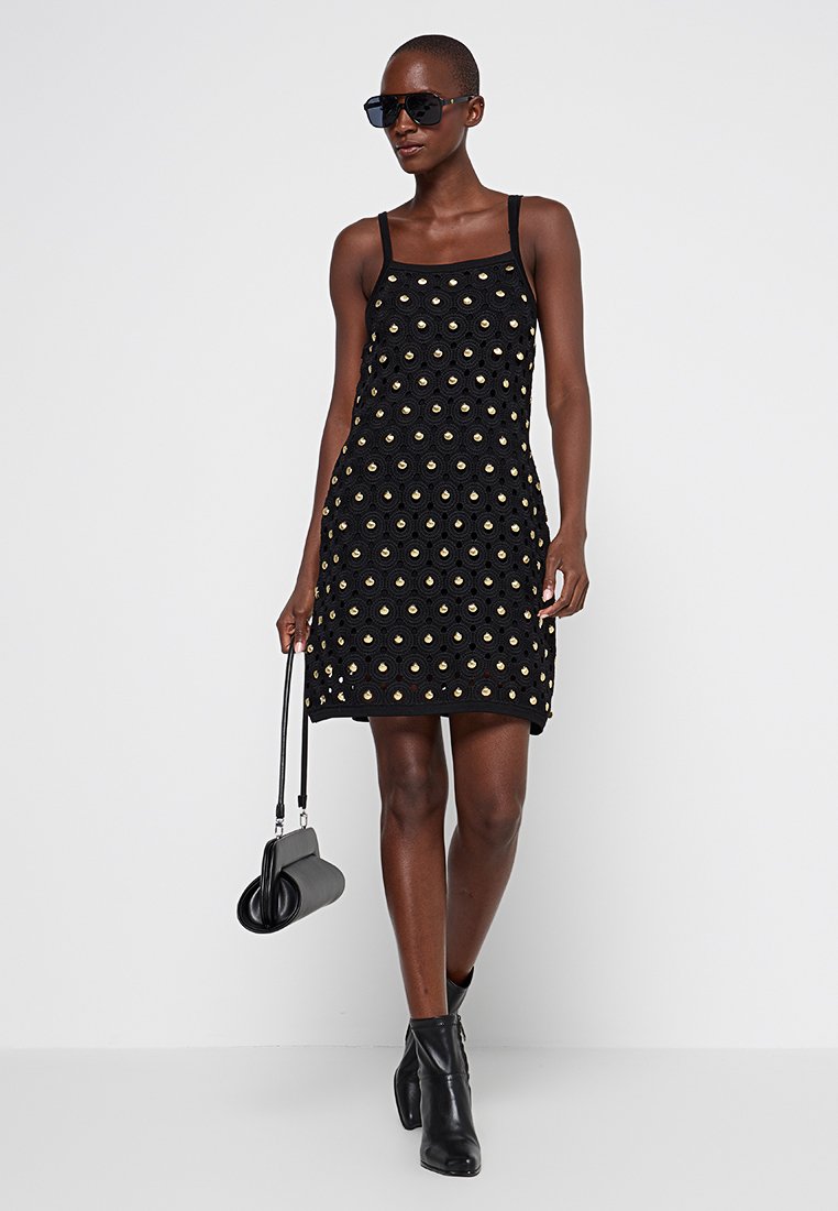 Frau in schwarzem ärmellosem Kleid mit kreisförmigen Ausschnitten, schwarzen Knöchelstiefeln, Sonnenbrille und einer kleinen schwarzen Handtasche.