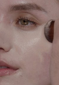 Das Make-up wird mit einem weichen Pinsel auf eine glatte, ebenmäßige Hautoberfläche aufgetragen. Die Haut erscheint leicht glänzend mit sichtbarer Wärme in den Farben.