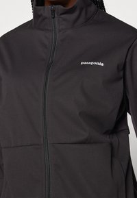 Giacca softshell nera con colletto alto, zip completa e logo Patagonia sul petto, caratterizzata da una texture liscia e opaca.