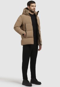 Bruine pufferjas met een capuchon, voorzien van een gewatteerd ontwerp, zwarte ritsen en zijzakken, gecombineerd met zwarte broek en laarzen.