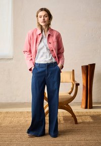 Femme debout, les mains dans les poches, portant une veste rose, un chemisier blanc, un pantalon large bleu, près d'une chaise en bois et d'une sculpture dans une pièce aux tons neutres.