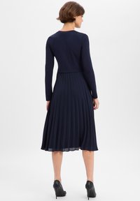 Kobieta ubrana w granatową bluzkę z długim rękawem i plisowaną spódnicę midi oraz czarne buty na wysokim obcasie, stojąca tyłem na białym tle.