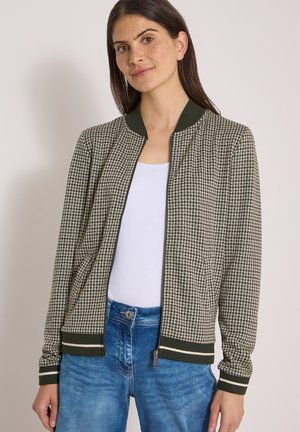 Femme portant une veste à fermeture éclair à motif pied-de-poule avec col et poignets côtelés vert foncé, au-dessus d’un haut blanc et d’un jean bleu.