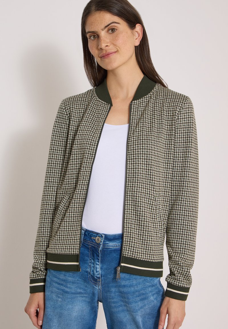 Femme portant une veste à fermeture éclair à motif pied-de-poule avec col et poignets côtelés vert foncé, au-dessus d’un haut blanc et d’un jean bleu.
