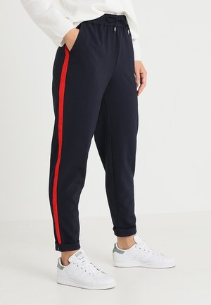 Person trägt marineblaue Jogginghosen mit roten Seitenstreifen und weiße Sneakers, Hände in den Taschen, steht vor einem einfarbigen hellen Hintergrund.