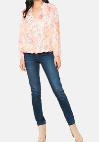 Kvinna som bär en pastellblommig långärmad blus, mörkblå skinny jeans och beige ankelstövlar, stående med ett ben korslagt.