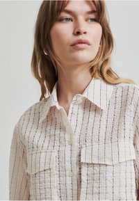 Camisa de crema texturizada con rayas verticales negras, dos bolsillos en el pecho y un cuello clásico con cierre de botón. Fabricada en un tejido suave y ligero.
