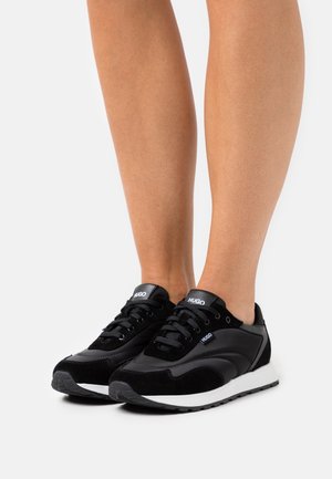 Jambes portant des baskets noires HUGO avec semelles blanches, dotées d'un design à lacets et d'étiquettes de marque sur la languette et le côté.