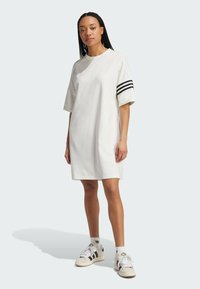 Abito t-shirt bianco oversize con maniche corte, caratterizzato da tre strisce nere su una manica, abbinato a sneakers bianche e calzini a fantasia.