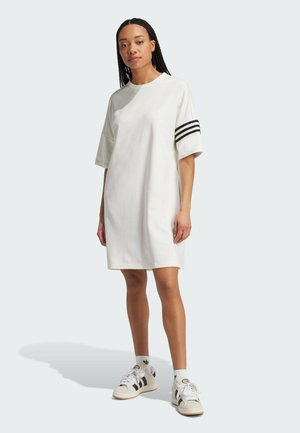 Abito t-shirt bianco oversize con maniche corte, caratterizzato da tre strisce nere su una manica, abbinato a sneakers bianche e calzini a fantasia.