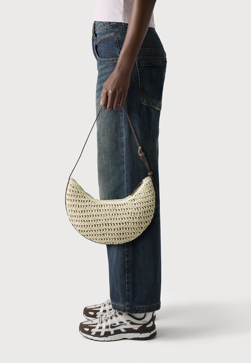 Crochet bag in cream with a half-moon shape, featuring a brown strap. Model wears blue jeans and white-brown sneakers.
