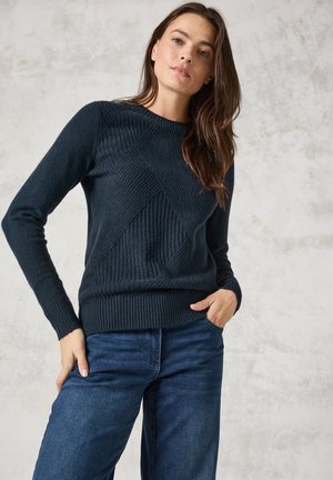 Granatowy sweter z dzianiny z teksturowanymi wzorami w diamenty, ze ściągaczami na mankietach i u dołu, w połączeniu z niebieskimi dżinsami. Miękki, luksusowy materiał.