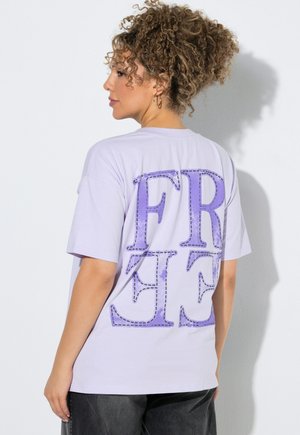 Frau mit lockigem Haar, die ein helllila T-Shirt mit großen violetten Buchstaben "FREE" auf dem Rücken trägt, steht an einer weißen Wand.