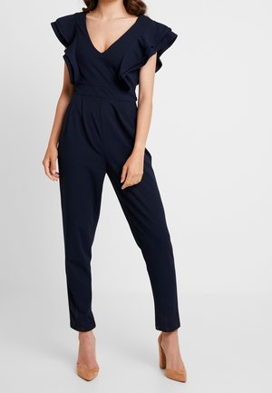 Kvinde iført en marineblå ærmeløs jumpsuit med flæser på skuldrene og brune høje hæle, stående mod en ensfarvet baggrund.