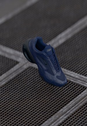 Chaussure de sport bleu foncé avec semelle noire suspendue au-dessus d'un sol en grille métallique texturée.