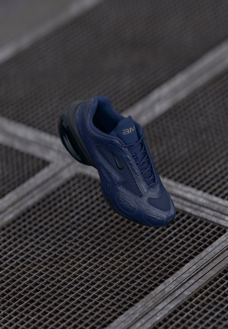 Chaussure de sport bleu foncé avec semelle noire suspendue au-dessus d'un sol en grille métallique texturée.