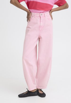 Persoană îmbrăcată în blugi roz cu talie înaltă și pantaloni largi, balerini negri și un top cu mâneci scurte cu dungi roșii și roz, cu mâinile pe șolduri.