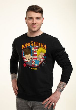 Mężczyzna w czarnej bluzie Beavis and Butt-Head z animowanymi postaciami i amerykańską flagą w tle, stoi z rękami na biodrach.