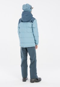 Hellblauer Puffer-Jacke mit einer dunkler blauen Kapuze und Schulter, graue Hose und schwarze Stiefel. Die Jacke hat ein gestepptes Design und elastische Bündchen.