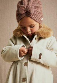 Hvid fleece frakke med pelskrave, træknapper og et brunt label. Et barn justerer frakken, iført en lyserød strikket buehue.