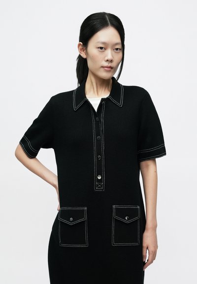 Fekete ruha gallérral, rövid ujjú, gombokkal az elején, és kontrasztos fehér varrással. Elöl két zseb található, zsebfedőkkel.