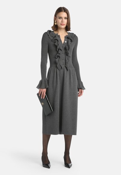 Abito in maglia grigia a coste con scollatura arricciata e chiusura con bottoni. Include maniche svasate, abbinato a una pochette nera e tacchi a punta.
