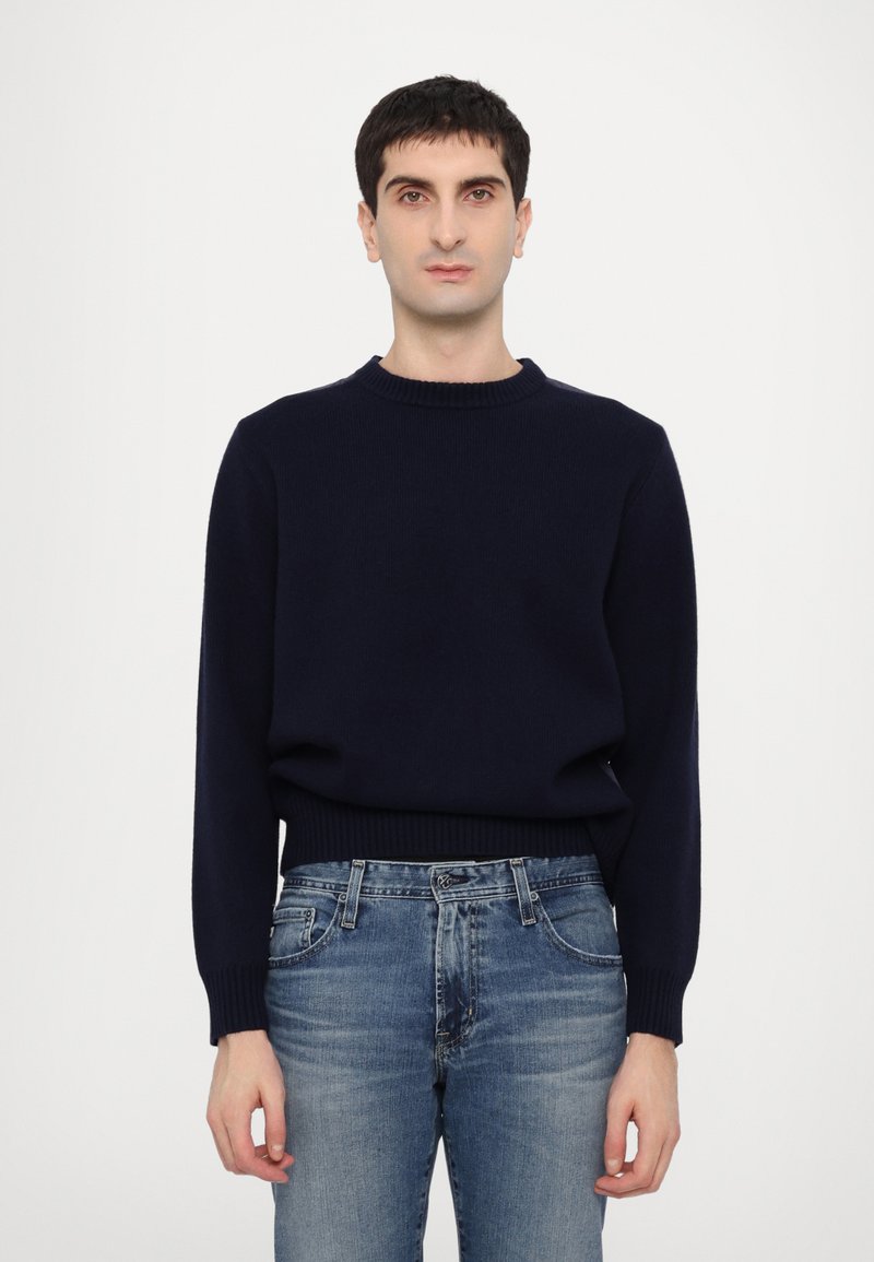 Marinblå stickad tröja med ribbad rund halsringning, långa ärmar och avslappnad passform, i kombination med blå jeans i en blekt denimtvätt.