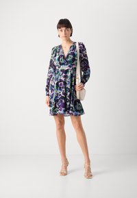 Robe à imprimé floral en violet, vert et bleu, avec un décolleté en V profond, des manches longues et une taille élastique. Portée avec un sac à bandoulière léger.