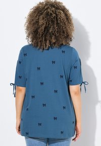 T-shirt en coton bleu à manches courtes, présentant un motif de petits nœuds noirs et des liens aux manches. Design à coupe décontractée, texture légère.