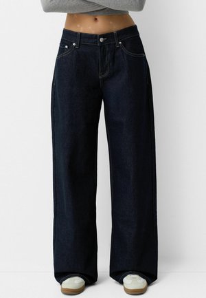 Personne portant un jean large foncé, des baskets blanches et un haut court gris à manches longues, debout les bras croisés.