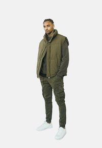Gilet rembourré vert sur un pull vert foncé, associé à un pantalon cargo olive et des baskets blanches. Texture notable dans le tissu ; design ajusté.