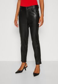 Žena v černých kožených kalhotách, černých lodičkách na podpatku a červeném topu, stojící na jednobarevném světlém pozadí.