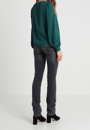 Kobieta ubrana w zieloną bluzkę z długim rękawem, ciemnoszare obcisłe dżinsy i czarne botki na obcasie stojąca na jednolitym tle.
