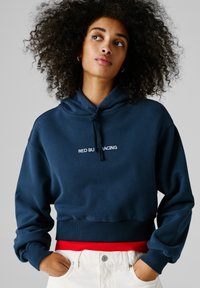 Žena s kučeravými vlasmi v tmavomodrej mikine s nápisom „RED BULL RACING“, bielych nohaviciach a rukami v vreckách na sivom pozadí.