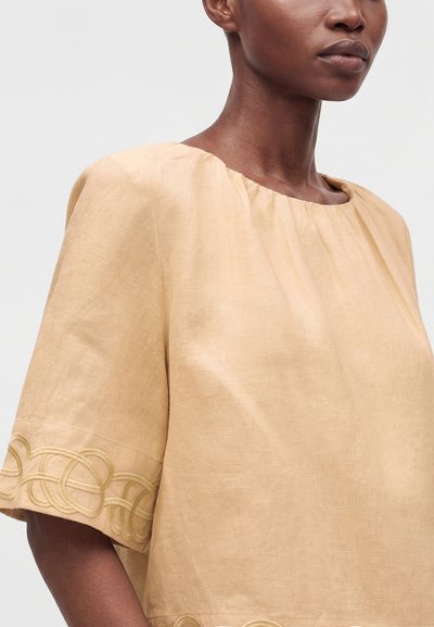 Personne à la peau foncée portant un haut beige ample à manches courtes avec des boucles décoratives cousues près des poignets et de l'ourlet.