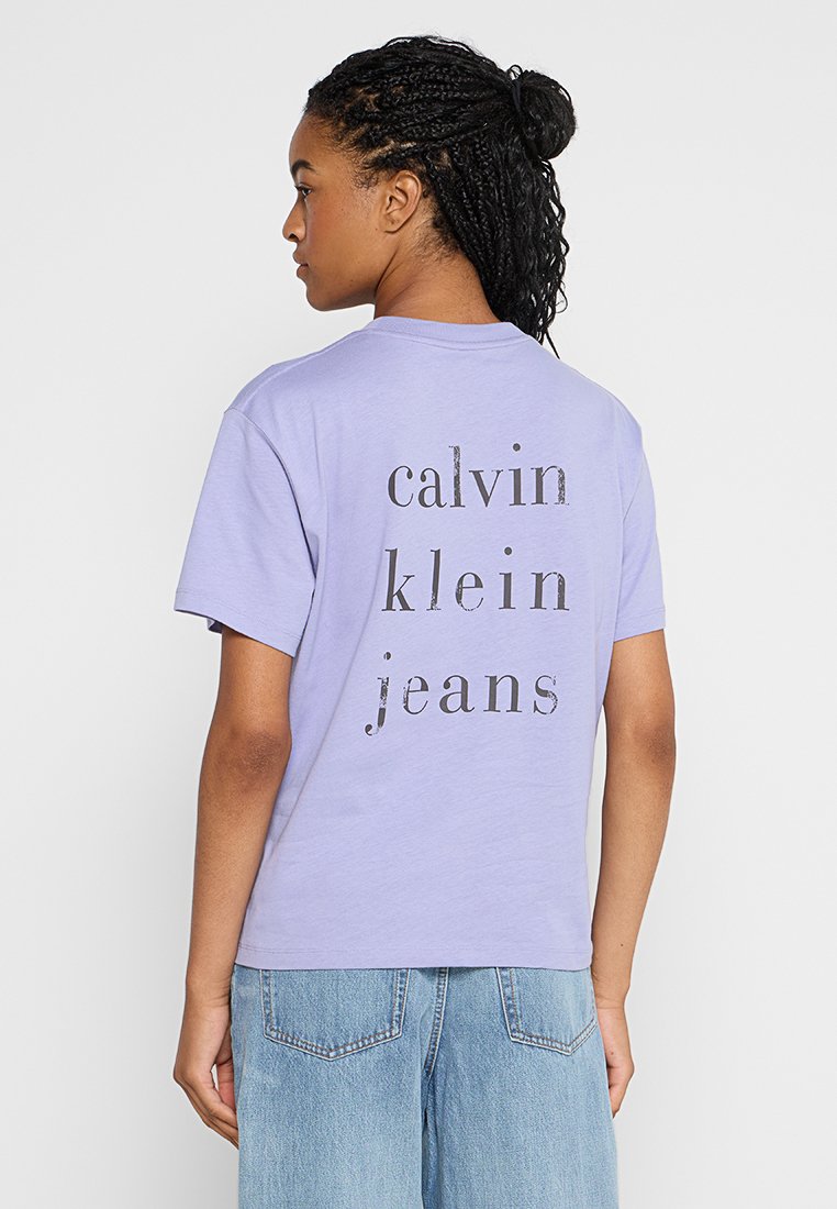 Kobieta z warkoczami, ubrana w fioletową koszulkę Calvin Klein Jeans i jasnoniebieskie dżinsy, tyłem do kamery.