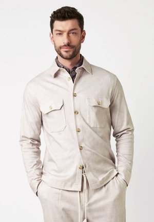 Hellbeige gestreifte Jacke mit zwei Brusttaschen und goldenen Knöpfen. Der Stoff hat eine weiche Textur und ist mit einem gemusterten Hemd darunter kombiniert.