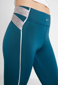 Turkusowe legginsy sportowe z szerokim szarym pasem wytłoczonym napisem "NIKE" oraz odblaskowym paskiem z boku dla lepszej widoczności. Gładka tekstura.