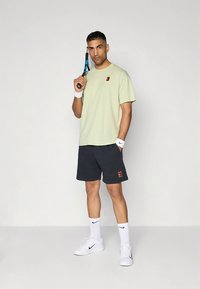 Man i tennisoutfit som håller ett racket över axeln, bär ljusgrön tröja, svarta shorts, vita Nike-strumpor, handledsband och sneakers.