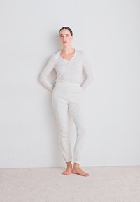Kvinna med mörkt hår i en låg knut, klädd i en vit långärmad henley-tröja och texturerade vita byxor, stående barfota mot en enkel vägg.