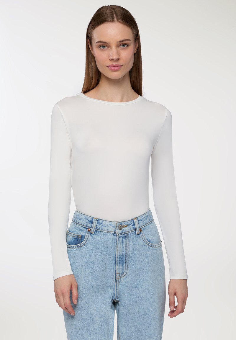 Maglietta bianca a maniche lunghe e aderente con scollo rotondo, abbinata a jeans in denim blu chiaro a vita alta con lavaggio sbiadito e cuciture visibili.