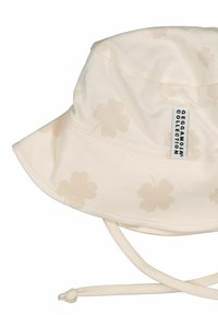 Kermanvärinen ämpärihattu, jossa on hillitty kukkakuosi, pehmeä tekstuuri ja säädettävä hihna tukevan istuvuuden takaamiseksi. Brändilogo näkyvissä.