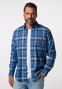 Camisa de cuadros azul con cuadrados de tonos oscuros y claros. Presenta un collar, mangas largas y detalles de botones. Lleva puesta una camiseta blanca debajo.
