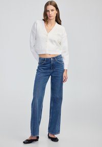 Kobieta ubrana w biały sweter zapinany na guziki, niebieskie dżinsy o prostym kroju i czarne płaskie buty, stoi na tle jednolitego jasnoszarego tła.