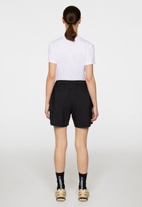 Svarta sportshorts med elastisk midja, kombinerat med en vit kortärmad skjorta. Modellen sedd bakifrån, iförd svarta strumpor och skor.