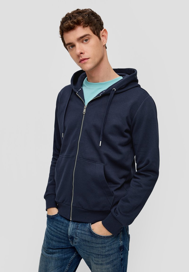 Sudadera con cremallera color marino, hecha de una suave mezcla de algodón, que cuenta con un bolsillo canguro, cordones y puños acanalados, llevada sobre una camiseta azul claro.