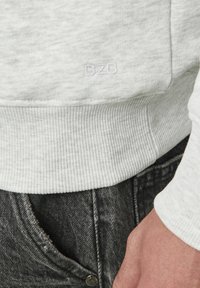 Sweatshirt gris clair avec des poignets et un ourlet côtelés, arborant un logo "B2B" en relief. Porté sur un tissu gris foncé avec une texture denim visible.