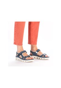 Blå och vita öppna sandaler med justerbara remmar, texturerad ovandel och vit vadderad sula, visade tillsammans med korallfärgade korta byxor.